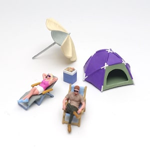 May include: A miniature camping set featuring a purple dome tent, a beige beach umbrella, a blue cooler, and two small figures. One figure wears a pink top and white shorts on a lounge chair, while the other wears a tan shirt on a folding chair.