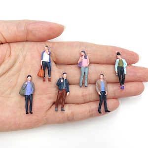 Puede incluir: Seis figuras de plástico en miniatura de personas con ropa informal, incluyendo varias camisas, chaquetas, pantalones y accesorios como bolsos, dispuestas en grupo. Cada modelo a pequeña escala presenta detalles pintados distintos.