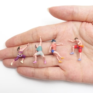 Puede incluir: Cuatro figuras en miniatura que representan personas en poses atléticas. La primera viste top y leggings morados, la segunda camiseta sin mangas verde azulado y pantalones cortos grises, la tercera camiseta coral y pantalones cortos azules, y la cuarta pantalones cortos rojos y cinturón amarillo.
