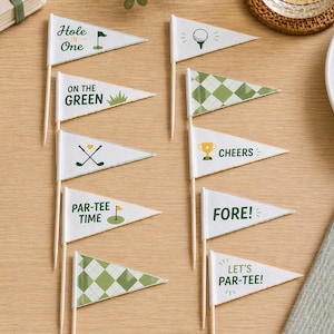 Può includere: Una collezione di bandierine triangolari bianche a tema golf con varie frasi e grafiche, tra cui "Hole One", "On the Green" e "Par-Tee Time". Le bandierine sono su bastoncini di legno, con una pallina da golf e biscotti visibili.