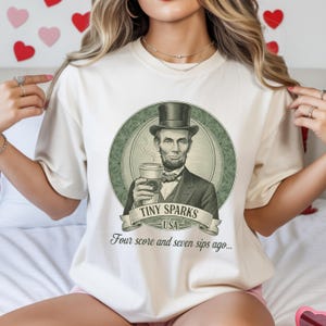 Puede incluir: Camiseta color crema con un gráfico de Abraham Lincoln sosteniendo una taza de café. El diseño incluye el texto "TINY SPARKS USA" y "Four score and seven sips ago...". La imagen está dentro de un marco circular verde.