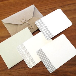 May include: A stationery set comprising a kraft paper envelope with a string closure and several white cards. Some cards have grey patterned borders, including a houndstooth and a plaid design. The set is arranged on a wooden surface.