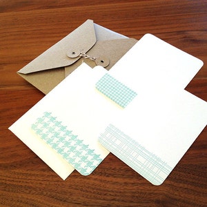 May include: Three white stationery cards with blue patterned borders. The cards are stacked on top of each other. The top card has a small blue patterned tag attached to it. The cards are on a brown wooden surface.