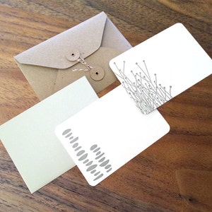 May include: Three blank note cards with different designs. One card has a gray pattern of dots, another has a gray pattern of lines, and the third card is blank. The cards are on a wooden surface.