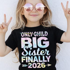 Peut inclure: Un t-shirt noir avec le texte "ONLY CHILD BIG Sister FINALLY 2026" dans un motif tie-dye coloré. La fille porte des lunettes de soleil rondes teintées de rose et fait le signe de la paix avec ses doigts.