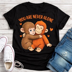 May include: Black t-shirt with the text "YOU ARE NEVER ALONE" in orange. The shirt features a cartoon illustration of two monkeys hugging, with orange hearts. White sneakers and denim shorts are visible.