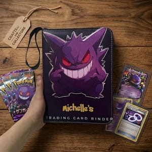 Könnte beinhalten: Ein schwarzer Sammelkartenordner mit einem großen lila Gengar-Charakter mit roten Augen und einem breiten Grinsen. Der Text "Michelle's" und "TRADING CARD BINDER" ist in Gelb und Weiß aufgedruckt. Enthält ein Etikett "CRAFTED FOR COLLECTORS".