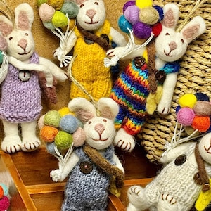 Puede incluir: Adornos de conejos hechos a mano. Cada conejo viste trajes de punto de varios colores y sostiene un montón de globos de fieltro de colores. Los conejos tienen pelaje blanco, orejas rosas y ojos de botón marrones. Se exhiben en una caja de madera.