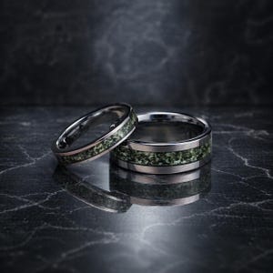 May include: Two silver rings with a green speckled inlay, set against a dark, reflective backdrop. The rings' design is mirrored in the surface below. These rings could be wedding bands or engagement rings.