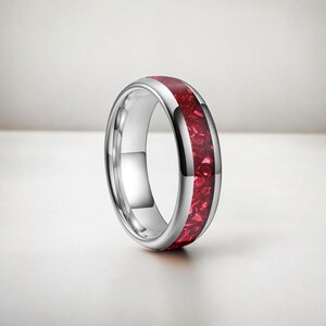 Könnte beinhalten: Ein silberner Ring mit einem leuchtend roten Einsatz. Der Ring hat eine polierte, reflektierende Oberfläche und ein Band aus zerkleinertem rotem Material. Das Design ist schlicht und modern, geeignet für den Alltag oder besondere Anlässe.