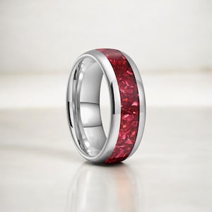 May include: A silver-colored ring with a wide band of vibrant red material. The ring has a polished, reflective surface, and the red material appears to be made of small, irregular pieces. The ring is a piece of jewelry.