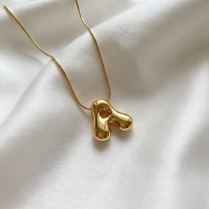Puede incluir: Un collar dorado con un colgante abstracto único. El colgante tiene una forma redondeada y orgánica con un agujero en el centro. La cadena es fina y dorada, sobre un fondo de satén blanco.