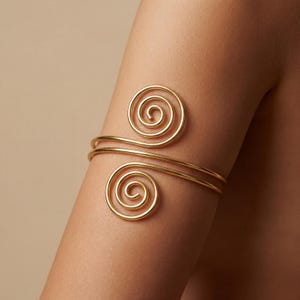 Puede incluir: Un brazalete dorado compuesto por una banda de metal lisa y pulida que se enrolla alrededor del brazo. El diseño termina en ambos extremos con una forma de espiral circular distintiva, creando una silueta abierta y ajustable.