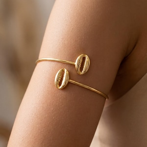 May include: A gold-toned open cuff bracelet featuring two polished cowrie shell charms at each end. The metal band is smooth and circular, designed to wrap around the arm, with the two shells positioned to face each other in a bypass style.