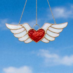May include: A stained glass heart with white wings and gold-colored trim, suspended by a gold chain. The heart is a vibrant red, and the wings have an iridescent sheen. The background is a bright blue sky with scattered clouds.