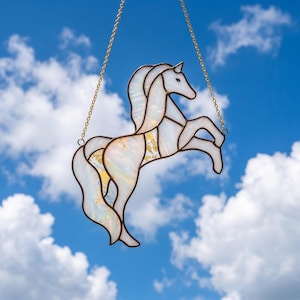 May include: A stained glass horse in a rearing pose, with a white and iridescent body and gold accents. The horse is outlined in dark brown and suspended by a gold chain against a blue sky with white clouds. A decorative piece.