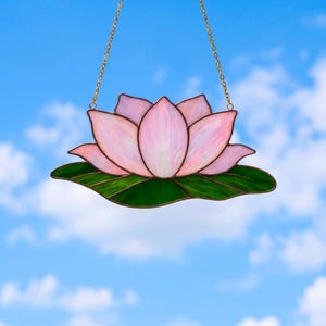 May include: A stained glass lotus flower with pink petals and green leaves, suspended by a gold chain. The flower is set against a blue sky with white clouds. The handcrafted piece is a decorative item.