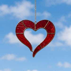 May include: A red stained glass heart-shaped suncatcher with a gold-coloured metal frame and hanging chain. The heart is divided into sections and hangs against a blue sky with white clouds.
