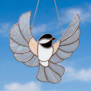 Puede incluir: Un pájaro de vidriera con las alas extendidas, con cabeza negra, pecho blanco y alas grises. El pájaro está suspendido por una cadena plateada contra un cielo azul. Las alas tienen una cualidad iridiscente.