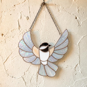 May include: A stained glass suncatcher shaped like a chickadee with wings spread wide. The bird features iridescent white and pale blue glass panels, a black head, and a white chest, all held together by copper-colored metal solder and a hanging chain.