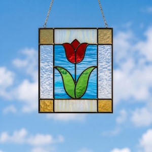 May include: Stained glass panel featuring a red tulip with green leaves against a blue background. The panel is framed in black with gold and white accents, and is suspended by a gold chain. The artwork is set against a bright blue sky.