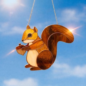 May include: A stained glass suncatcher shaped like a squirrel holding an acorn. The squirrel is crafted from various shades of amber, brown, and cream glass pieces joined by metal solder lines, suspended by a thin silver-toned metal chain.