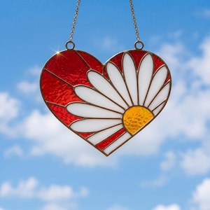 May include: Stained glass heart-shaped suncatcher with a daisy design. The heart features red and white glass petals with a yellow center. The suncatcher hangs from a gold-colored chain against a blue sky.