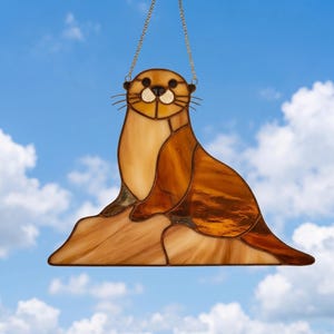 Pode incluir: Um apanhador de sol de vitral em forma de lontra sentada numa rocha, com peças de vidro em vários tons de castanho e bronze unidas por linhas de metal escuro, suspenso por uma fina corrente de metal dourado para pendurar na janela.