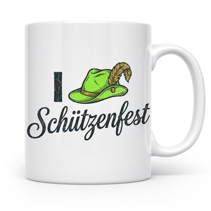 Könnte beinhalten: Weiße Keramik-Tasse mit dem Text "I Schützenfest" in schwarzer Schrift. Das "I" ist ein schwarzer vertikaler Strich, und der Text "Schützenfest" befindet sich unter einem grünen Hut mit einer Feder.
