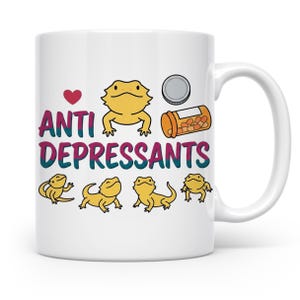 Könnte beinhalten: Ein weißer Keramikbecher mit einem roten Herz, fünf gelben Bartagamen-Illustrationen, einer Pillendose und dem Text "ANTI DEPRESSANTS" in Lila und Türkis. Das Design enthält einen kleinen grauen runden Deckel neben der orangefarbenen Dose.