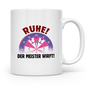 Könnte beinhalten: Weißer Keramikbecher mit einem Dartscheiben-Design in Pink und Lila, mit drei Darts in der Mitte. Der Becher trägt den Text "RUHE!" über der Dartscheibe und "DER MEISTER WIRFT!" darunter.