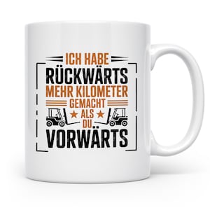 Könnte beinhalten: Weiße Keramik-Tasse mit schwarzer und brauner Grafik. Der Text auf der Tasse lautet "ICH HABE RÜCKWÄRTS MEHR KILOMETER GEMACHT ALS DU.. VORWÄRTS" mit zwei Gabelstapler-Illustrationen. Die Tasse hat einen weißen Henkel.