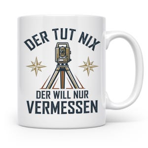 Könnte beinhalten: Ein weißer Keramikbecher mit einer Grafik eines Tachymeters auf einem Stativ, flankiert von zwei goldfarbenen Kompasssternen. Der Text "DER TUT NIX" steht über dem Gerät und "DER WILL NUR VERMESSEN" in dunkelblauer Schrift darunter.