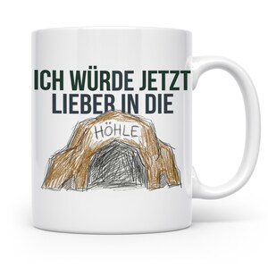 Könnte beinhalten: Ein weißer Keramikbecher mit einem dunkelgrünen Textaufdruck "ICH WÜRDE JETZT LIEBER IN DIE" über einer handgezeichneten Illustration einer braunen Höhle, bei der das Wort "HÖHLE" in schwarzer Tinte am Eingang der Höhle zu lesen ist.