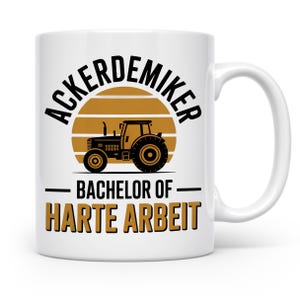 Könnte beinhalten: Weiße Keramiktasse mit einer schwarzen Traktorgrafik vor einem braunen Sonnenuntergangsdesign. Die Tasse trägt den Text "ACKERDEMIKER" über dem Traktor und "BACHELOR OF HARTE ARBEIT" darunter.