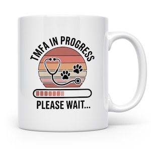 Könnte beinhalten: Weißer Keramikbecher mit schwarzem Henkel. Der Becher zeigt den Text "TMFA IN PROGRESS" in einem kreisförmigen Design, ein Stethoskop, Pfotenabdrücke und eine Ladeleiste mit "PLEASE WAIT..."