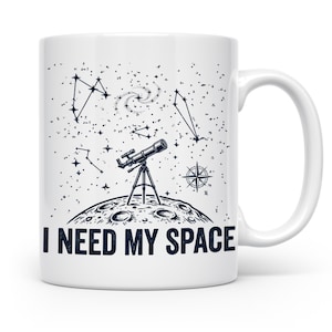 Könnte beinhalten: Weiße Keramik-Tasse mit einer schwarzen Illustration eines Teleskops auf dem Mond, Sternbildern und dem Text "I NEED MY SPACE". Die Tasse hat einen weißen Henkel und ist für Heißgetränke konzipiert.
