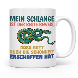 Könnte beinhalten: Ein weißer Keramikbecher mit einer Grafik einer grün-gelb gemusterten Schlange, die zu einer Schleife gewunden ist. Der Text lautet: MEIN SCHLANGE IST DER BESTE BEWEIS, DASS GOTT AUCH DIE SCHÖNHEIT ERSCHAFFEN HAT. Der Becher hat einen Henkel.