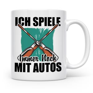 Könnte beinhalten: Weiße Keramik-Tasse mit dem schwarzen Text "ICH SPIELE IMMER NOCH MIT AUTOS" und gekreuzten Schraubendrehern. Das Design beinhaltet einen türkisgrünen Hintergrund. Die Tasse hat einen weißen Henkel.