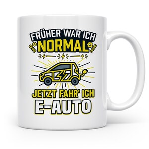 Könnte beinhalten: Ein weißer Keramikbecher mit einer Grafik eines gelben Elektroautos mit Blitzsymbolen. Der Text lautet "FRÜHER WAR ICH NORMAL" in Schwarz und Gelb sowie "JETZT FAHR' ICH E-AUTO" in Schwarz und Gelb, umgeben von kleinen Zahnradsymbolen.