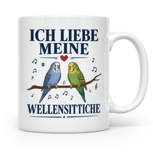 Könnte beinhalten: Eine weiße Keramiktasse mit einem blauen und einem grünen Wellensittich auf einem braunen Ast. Der Text lautet ICH LIEBE MEINE WELLENSITTICHE in dunkelblauen Buchstaben, ergänzt durch ein kleines rotes Herz und Notensymbole.