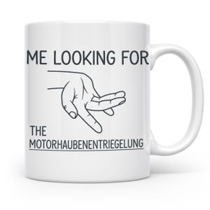 Könnte beinhalten: Eine weiße Keramiktasse mit einer schwarzen Strichzeichnung einer Hand, die nach etwas sucht. Der aufgedruckte Text lautet ME LOOKING FOR THE MOTORHAUBENENTRIEGELUNG in schwarzen Großbuchstaben, wobei das letzte Wort unterstrichen ist.