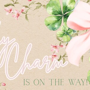 May include: A light beige background with the text "A Little Lucky Charm Is On The Way!" in a cursive font. The image features a large pink bow, green shamrocks, and pink flowers.