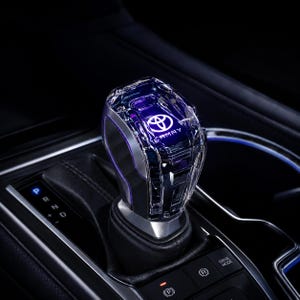 May include: A crystal-style gear shift knob featuring a clear, faceted exterior with an illuminated Toyota logo and the word CAMRY visible inside. The knob is mounted on a black leather-wrapped base within a vehicle centre console.