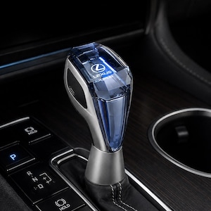 May include: A Lexus gear shift knob featuring a translucent blue crystal-like top section with the Lexus logo clearly visible inside. The base is constructed from a matte silver metallic material, mounted on a black leather boot with white stitching.