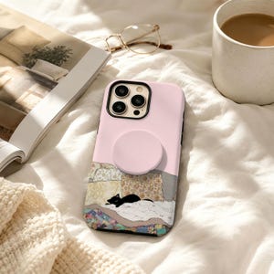 May include: A light pink smartphone case featuring a circular grip attachment on the back. The lower portion of the case displays a graphic illustration of a small black cat resting on a colorful patchwork quilt with various floral and geometric patterns.