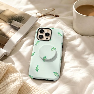 May include: A light mint green smartphone case featuring a repeating pattern of small, bright green geckos. The case includes a circular, matching mint green collapsible grip attached to the centre back and a black camera lens frame.