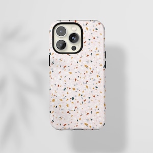 Puede incluir: Una funda de teléfono con un patrón de terrazo. La funda tiene un fondo rosa claro con pequeñas motas de colores azul, amarillo, marrón y rojo. El área de la cámara es negra y la funda tiene un borde negro.