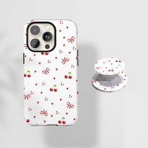 May include: White phone case and matching phone grip featuring a repeating pattern of red cherries, bows, and hearts. The phone case has a black camera ring and black side buttons. The phone grip is round and has a white base.