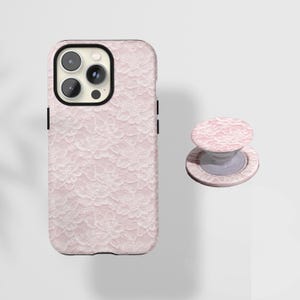May include: A pink phone case and matching phone grip featuring a delicate white lace pattern. The phone case has a black camera border and black side buttons. The phone grip is round and has a collapsible design.